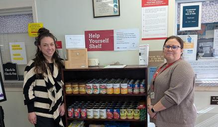 Two women standing next to a shelf stocked with non-perishable food items 