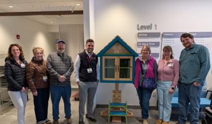 A group of adults standing together smiling next to a small house-shaped box containing books. They are celebrating together.