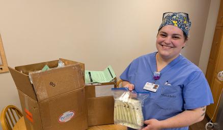 A woman in scrubs smiling and holding bags of donated breast milk standing next to at table with a large brown box on it