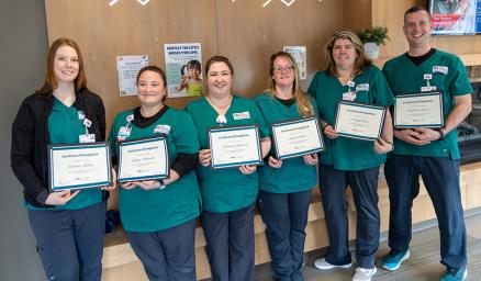 A group of adults wearing matching teal scrubs, standing together in a hallway holding up diplomas and smiling
