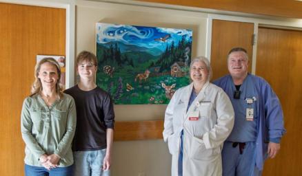 Four adults standing together and smiling next to a painting hanging on a hospital wall. The painting is of a peaceful woodland scene with bright green grass and blue skies