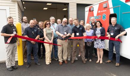 a group of adults participating in a ribbon cutting ceremony outside a building and next to an ambulance