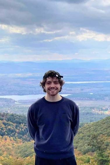 Michael Rowe smiling and standing atop a mountain with a large valley behind him covered by a thick cloudy sky
