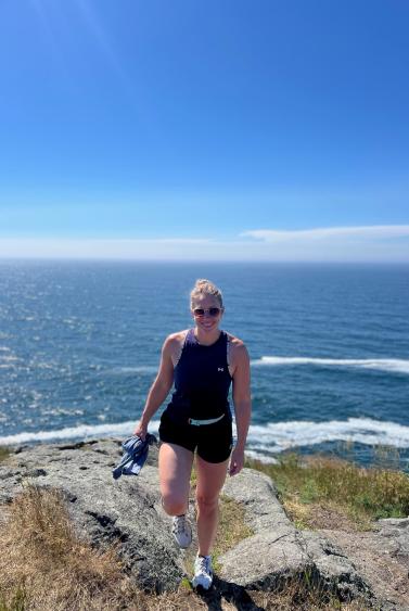 Sarah Carter standing on top of a rocky cliff overlooking the ocean that is behind her. She is looking straight ahead with a smile.