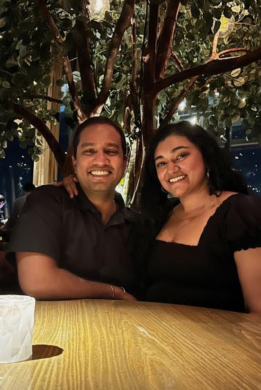 Two adults sitting together a dining table at night. A large tree is behind them and the area is lit by soft light. Both are smiling.