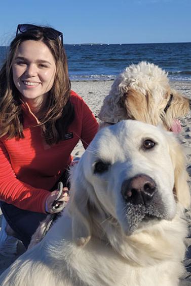 smiling young woman, crouched next to two light-colored dogs at the beach