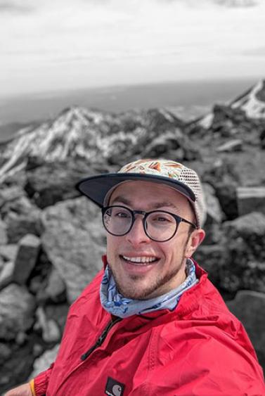 Young man in a white hat, glasses, and red jacket in front of a rocky coastline