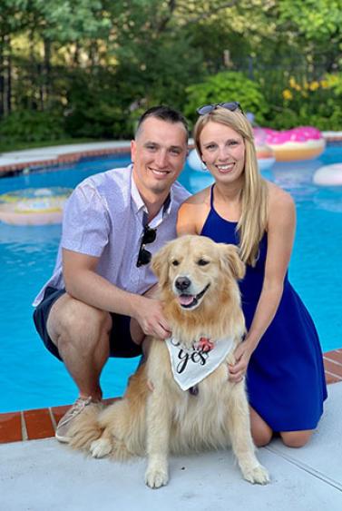smiling young man and woman crouched next to light-colored dog in front of a blue pool