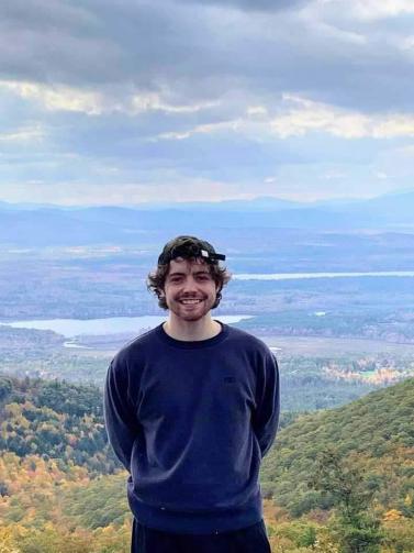 Michael Rowe smiling and standing atop a mountain with a large valley behind him covered by a thick cloudy sky