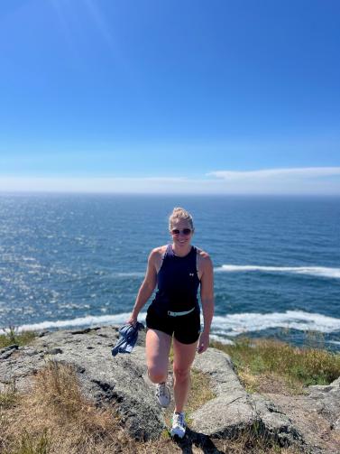 Sarah Carter standing on top of a rocky cliff overlooking the ocean that is behind her. She is looking straight ahead with a smile.