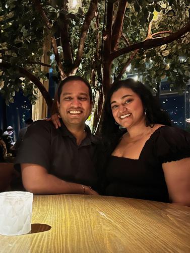Two adults sitting together a dining table at night. A large tree is behind them and the area is lit by soft light. Both are smiling.